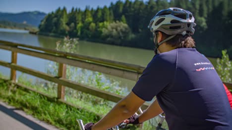 Two-happy-friends-cycling-along-the-river,-idyllic-view-of-forest-and-mountain