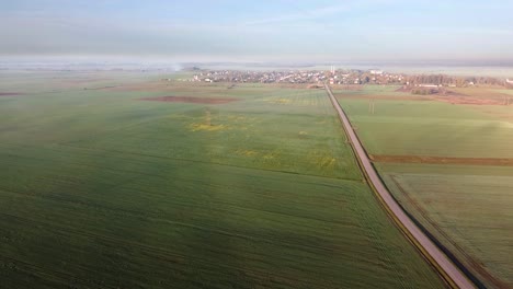 Drohne-Fliegt-Am-Frühen-Nebligen-Morgen-Mit-Sonnenaufgang-über-Einer-Kleinen-Landstraße-Mit-Grünen-Feldern