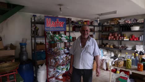 Old-Caucasian-Lebanese-man-in-small-food-marketplace-store-walks-towards-camera-and-waves-hello,-Douma,-Lebanon,-close-up-pan