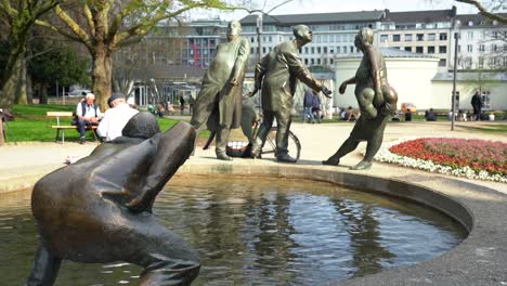 Estatua-De-Bronce-Con-Gente-Y-Piscina-De-Hidromasaje-En-Aquisgrán,-Alemania,-Llamada-Circulación-De-Dinero-Con-El-Elisenbrunnen-En-Segundo-Plano