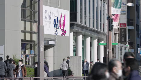 Personas-Con-Mascarillas-Caminando-En-La-Calle-En-Un-Día-Soleado-Con-La-Pancarta-Olímpica-Cancelada-De-Tokio-2020-En-El-Exterior-Del-Edificio-Durante-La-Pandemia-En-Tokio,-Japón