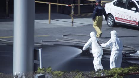 Un-Grupo-De-Hombres-Con-Trajes-De-Riesgo-Biológico-Limpian-El-Suelo-De-Una-Gasolinera-Con-Un-Chorro-De-Agua-Procedente-De-Una-Manguera-De-Bomberos-Durante-La-Pandemia-De-Covid-19