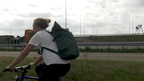 Una-Mujer-Joven-En-Bicicleta-Cerca-De-Una-Carretera-Principal,-En-Una-Zona-Urbana