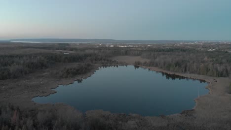 High-aerial-flight-over-a-blue-pond-in-the-evening-just-outside-of-Ottawa,-Ontario-with-the-city-skyline-in-the-distance