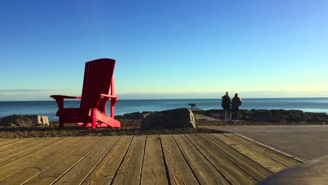 Wide-shot-of-beautiful-sunny-day-at-the-lake,-by-the-boardwalk,-with-oversized-red-cottage-chair-in-foreground-and-female-friends-having-a-leisurely-stroll-with-their-dog-in-the-background