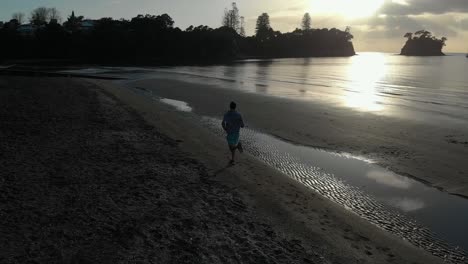 Aerial-follow-shot-of-silhouetted-young-man-running-on-a-beach-in-Auckland,-New-Zealand