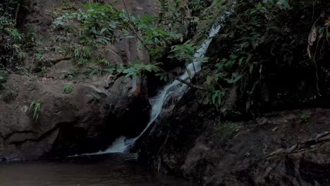 Sideways-sliding-movement-showing-a-waterfall-in-the-tropical-mountain-forest-of-Rio-de-Janeiro-mouthing-into-a-small-pond-and-rocky-surroundings