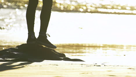 Las-Piernas-De-Una-Mujer-Con-Pantalones-Negros-Delgados-Y-Zapatos-Converse-Saliendo-De-Una-Pequeña-Roca-Hacia-La-Playa-De-Arena-Durante-Una-Brillante-Puesta-De-Sol-Dorada-Con-El-Océano-Al-Fondo