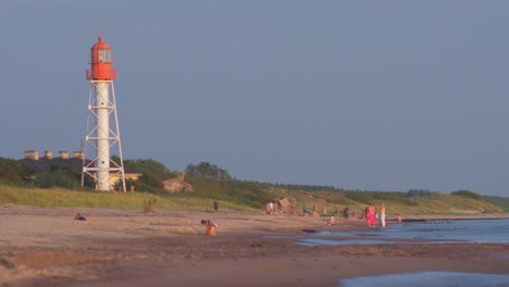 Die-Menschen-Genießen-Einen-Warmen-Sommerabend-Am-Strand-Mit-Einem-Leuchtturm-Im-Hintergrund-Vor-Sonnenuntergang