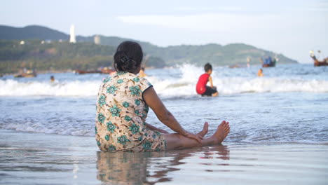 Mujer-Sentada-En-La-Orilla-De-La-Playa-Viendo-A-Sus-Hijos-Jugar-En-El-Océano