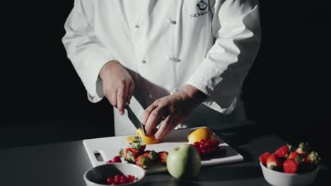 Cooking-Chef-Cuts-Slice-of-Lemon-on-Black-Table-Filled-with-Fresh-Fruits
