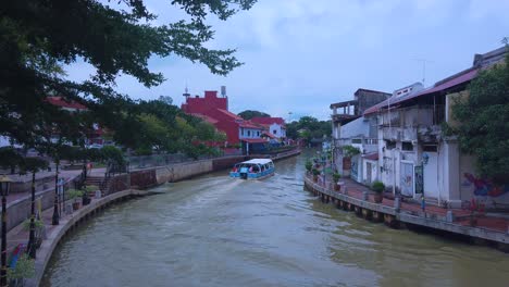 Barco-Turístico-Navegando-En-Un-Estrecho-Río-Melaka-Con-Gente-En-El-Barco,-Casas-Y-árboles-A-Ambos-Lados-Del-Río