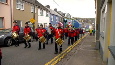 Banda-De-Músicos-Con-Banderas-Irlandesas-Con-Instrumentos-En-Las-Manos-Camina-Por-La-Calle-En-Un-Pequeño-Pueblo-Dingle-En-Una-Parte-Remota-De-Irlanda-En-El-Desfile-Del-Día-De-San-Patricio,-Típicas-Casas-Irlandesas-En-Segundo-Plano