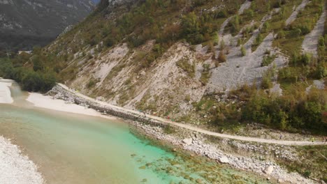 Wide-aerial-shot-of-marathon-runners-competing-at-marathon,-running-on-trail-path-next-to-emerald-Soca-river,-drone-moves-backwards,-late-summer-in-Soca-valley