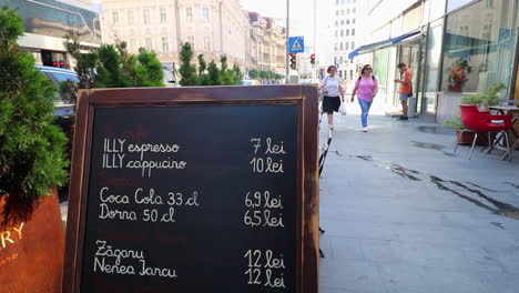 Women-walk-down-sunny-city-sidewalk-past-cafe-in-Bucharest,-Romania