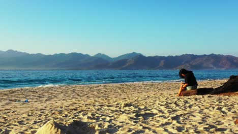 Mädchen-Sitzen-Auf-Einem-Getrockneten-Baumstamm-über-Dem-Weißen-Sand-Eines-Exotischen-Strandes-Und-Beobachten-Das-Blaue-Meer-Am-Klaren,-Hellen-Himmel-In-Bali