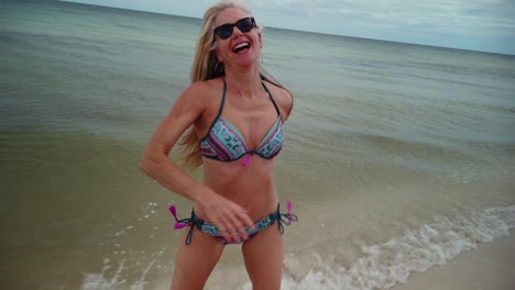 Wide-angle-view-of-laughing,-playful-mature-woman-in-bikini-and-sunglasses-spins-around-and-runs-on-the-beach