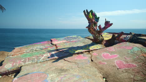 Graffiti-Art-on-Rocks-Overlooking-Bright-Blue-Pacific-Ocean-on-Sunny-Day,-Hearts-and-Painted-Tree