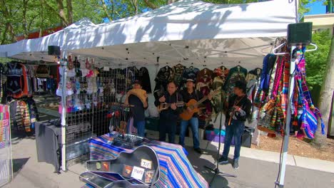 Four-musicians-who-are-signing-and-playing-a-wooden-pan-flute,-ukulele,-guitar-and-bells-at-an-open-air-market-in-the-park
