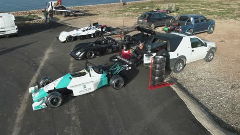 Hombre-Sentado-Cerca-Del-Coche-De-Carreras-Estacionado-En-El-Lado-De-La-Carretera-En-La-Colina-En-Imtahleb-Malta-Con-Un-Montón-De-Ruedas-Para-La-Reserva---Toma-Aérea