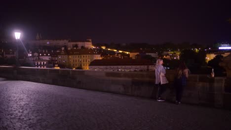 Two-girls-enjoying-a-nightly-stroll-and-watching-the-city-of-Prague-on-top-of-charles-bridge,-slow-pan-left,-young-tourists-in-Prague-at-night
