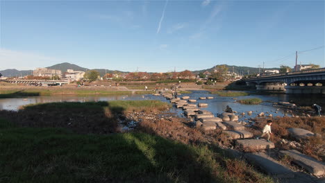 A-beautiful-steady-wide-shot-of-tourists-walking-on-rocky-pathway-on-Kamo-River-at-Kyoto,-Japan-during-afternoon-in-Autumn