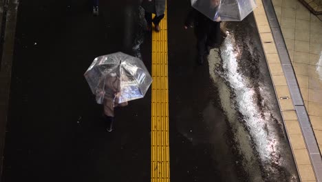 Top-down-shot-of-pedestrians-with-umbrellas-passing-below-a-bridge-on-a-rainy-night-in-Shinjuku,-Tokyo