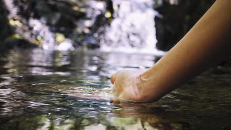 single-female-hand-touching-water-surface-at-waterfall,-slowmotion