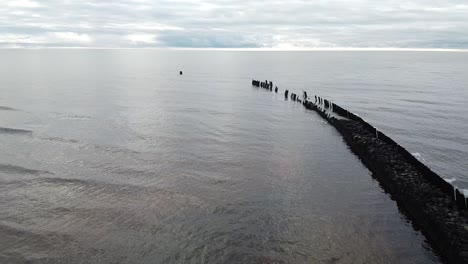 Aerial-view-of-old-breakwater---stone-pier-at-Pape-beach-Baltic-sea,-Latvia,-cloudy-and-calm-day,-wide-angle-establishing-drone-shot-moving-backwards