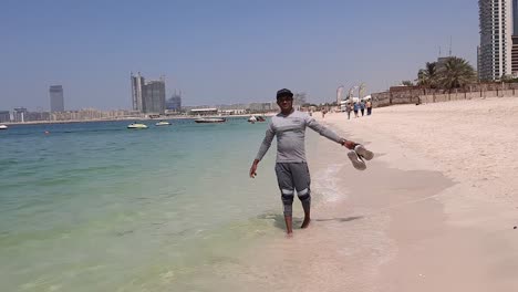 Un-Hombre-Asiático-Adulto-Camina-Por-La-Playa-Y-Se-Da-La-Vuelta,-Con-Las-Manos-Levantadas-Y-Abiertas-Felizmente-Y-Se-Aleja-De-Nuevo-En-La-Playa-De-Jumeirah-Día-Soleado