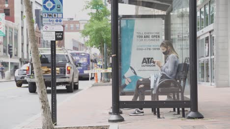 Frau-Sitzt-Auf-Einer-Bank-Im-Gläsernen-Busbahnhof,-Schaut-Aufs-Telefon-Und-Trägt-Gesichtsmaske