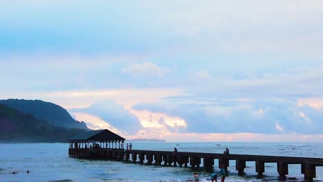 HD-Hawaii-Kauai-slow-motion-of-people-on-beach-with-Hanalei-Pier-in-midground-with-a-mountainous-coastline-in-distance-with-a-beautiful-colorful-sky-near-sunset,-also-available-in-4K-and-timelapse