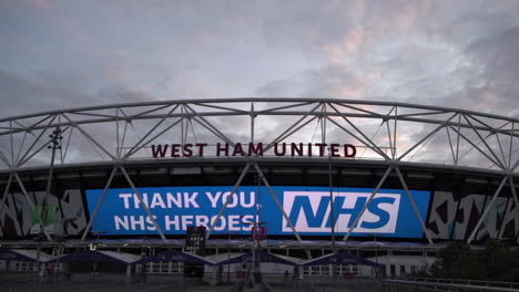 A-time-lapse-of-clouds-passing-over-the-Olympic-Park-Queen-Elizabeth-Stadium-at-sunset,-where-a-giant-billboard-praises-NHS-healthcare-workers-and-warns-the-public-to-maintain-social-distancing