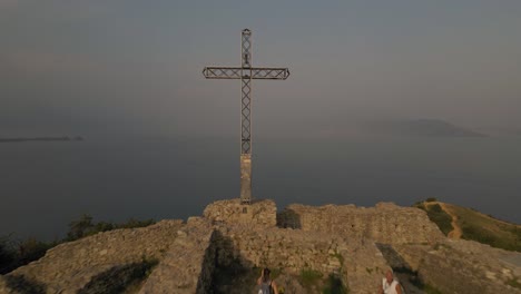 Lake-Garda-waters-in-mist-and-Rocca-di-Manerba-top,-cross-on-the-ruins,-aerial