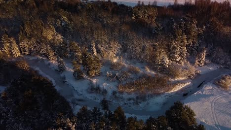 Beautiful-aerial-birdseye-view-of-snow-covered-fields-and-forest-near-frozen-Sventaja-river-in-sunny-winter-day,-golden-hour,-wide-angle-drone-shot-moving-backwards,-camera-tilt-down