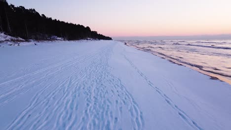 Beautiful-romantic-aerial-shot-flying-over-a-snow-covered-beach-after-the-sunset-on-the-calm-Baltic-sea-coastline,-high-contrast-sky,-wide-angle-drone-shot-moving-fast-forward