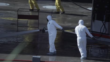 Un-Grupo-De-Hombres-Con-Trajes-De-Riesgo-Biológico-Limpian-El-Suelo-De-Una-Gasolinera-Con-Un-Chorro-De-Agua-Procedente-De-Una-Manguera-De-Bomberos-Durante-La-Pandemia-De-Covid-19