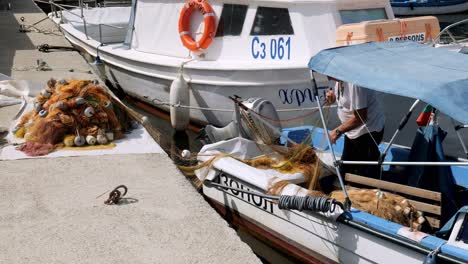 Man-mending-orange-fishing-nets-on-moored-boat-in-sozopol-marina-harbour