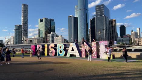 Wide-shot-of-people-having-fun-climbing-up-block-sign-of-Brisbane-and-taking-photos-of-iconic-landmark-with-downtown-cityscape-and-victoria-bridge-in-the-background,-Queensland,-Australia