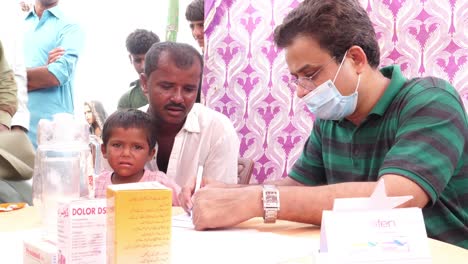 Trabajador-Humanitario-Con-Máscara-Facial-Anotando-Detalles-De-Los-Refugiados-De-Inundaciones-Con-Niños-En-El-Campamento-De-Socorro-En-Sindh