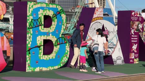 Family-and-tourists-taking-photos-with-giant-colorful-block-letter-of-Brisbane,-the-iconic-touristic-landmark-at-downtown-river-bank-on-a-sunny-afternoon,-Queensland-the-sunshine-state,-close-up-shot