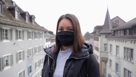 Attractive-female-model-with-respirator-mask-against-virus-infection-stands-in-front-of-city-of-Rapperswil,-Switzerland