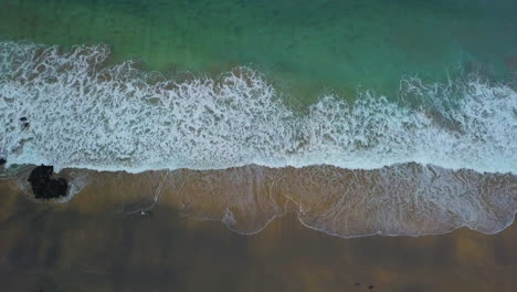 Top-down-aerial-view-of-waves-coming-up-onto-beach-as-people-walk-around