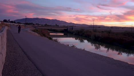 Cyclist-bikes-down-a-path-along-a-river-as-a-brilliant-sunset-reflects-off-the-water