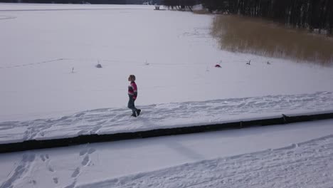 Slight-zoom-out-aerial-shot-of-young-woman-walking-on-snow-covered-jetty-on-edge-of-frozen-lake
