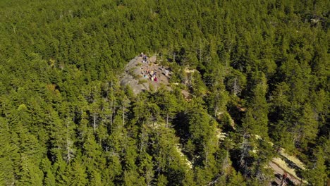 Aerial-drone-shot-over-the-green-and-autumn-colored-forest-and-a-group-of-hikers-on-a-rocky-outcrop-as-summer-ends-and-the-season-changes-to-fall-in-Maine