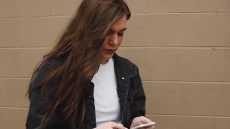 Steadicam-shot-of-a-beautiful,-brunette-college-teenager-texting-on-a-cell-phone-wearing-a-white-shirt-with-a-black-jacket-against-a-wall