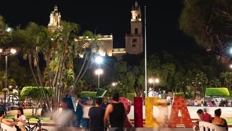 Time-lapse-with-motion-blurs-of-people-taking-their-photos-in-front-of-the-Merida-sign-in-the-Plaza-Grande-in-Merida,-Yucatan,-Mexico-at-night