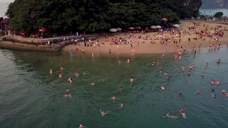 Toma-Aérea,-Dando-Vueltas-Sobre-Los-Nadadores-En-La-Excursión-A-La-Playa-En-El-Crucero-Por-La-Bahía-De-Ha-Long-A-última-Hora-De-La-Tarde,-Inclínate-Hacia-Arriba-Al-Final-Para-Revelar-El-Alto-Acantilado-De-La-Isla