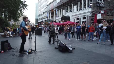 Singing-busker-on-Leicester-Square-with-crowd-and-person-donating-money-in-to-his-guitar-case,-London,-UK